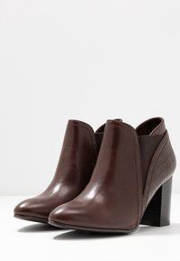 Botas de cuero marrón hasta el tobillo con un acabado brillante, paneles laterales elásticos, puntera puntiaguda y un panel trasero texturizado con patrón de cocodrilo. Tacón de altura media.