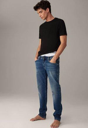 Schwarzes T-Shirt mit kurzen Ärmeln, kombiniert mit blauen Jeans aus Denim. Die Jeans haben eine klassische Passform, ausgebleichte Stellen und einen weißen Bund mit Logodetail.