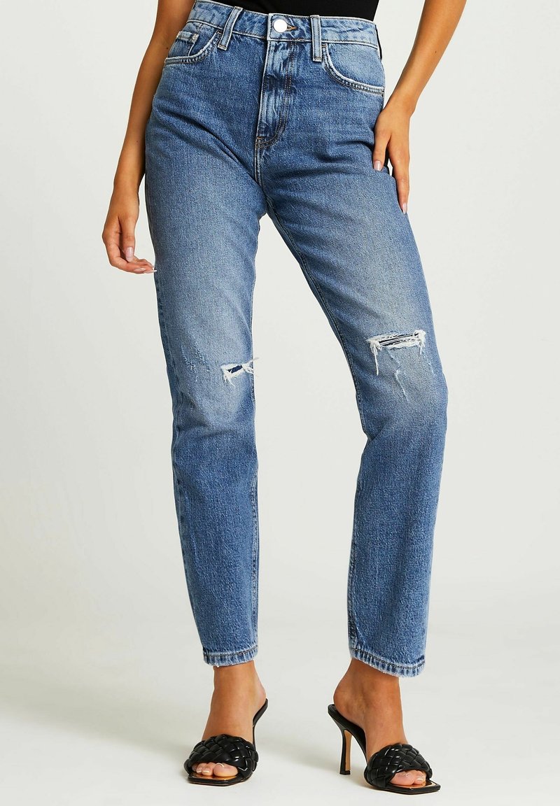 Höga blå jeans med slitna detaljer, rak passform och subtila blekningar. Kombinerat med svarta slip-on klacksandaler.