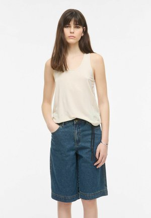 Ung kvinde med langt mørkt hår iført en ærmeløs beige top og løse denimshorts, der går til knæene, med den ene hånd i lommen.