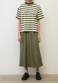 Chemise à rayures vertes et blanches à manches courtes, associée à une jupe verte unie de longueur mi-mollet. Le mannequin porte des chaussures noires à enfiler à semelle épaisse.