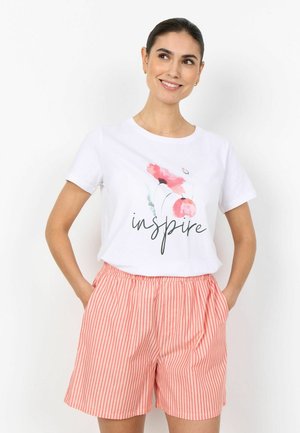 Lächelnde Frau mit dunklen Haaren, die ein weißes "inspire"-T-Shirt mit pinken Blumen trägt, dazu rosa-weiß gestreifte Shorts und die Hände in den Taschen hat.