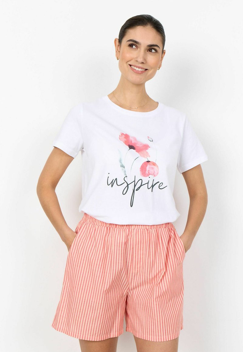 Femme souriante aux cheveux foncés portant un t-shirt blanc « inspire » avec des fleurs roses, un short rayé rose et blanc, les mains dans les poches.