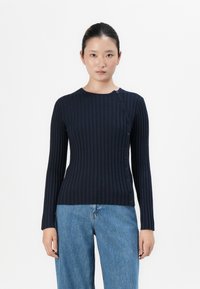 Navy-blauer, gerippter Strickpullover mit langen Ärmeln, Rundhalsausschnitt und seitlichem Knopfverschluss. Kombiniert mit hellblauen, lockeren Denim-Jeans.