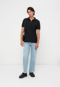 Melna polo krekla ar rāvējslēdzēju, gaiši zilas taisnas kājas džinsi un melni sporta apavi. Modelis stāv ar melnu plecu somu pret baltu fona.