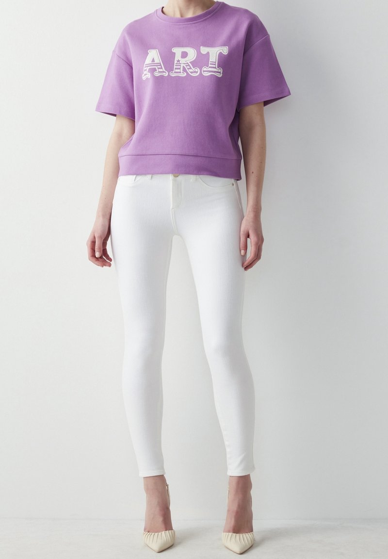 Lila cropped sweatshirt med "ART" tryck, kombinerad med vita skinny jeans och off-white klackar. Slät tyg och figurnära design.