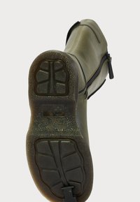 Semelle d'une botte en cuir vert avec un profil de caoutchouc noir texturé et une fermeture éclair sur le côté, inclinée vers le haut sur un fond blanc.