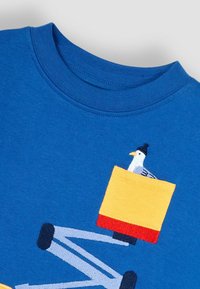 Kék, rövid ujjú póló, amelyen egy sárga zseb található, hímzett sirállyal, szürke és fekete csíkozású ujjakkal és bordázott gallérral.