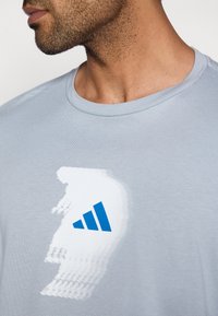 Light blue cotton t-shirt featuring a blurred white graphic and blue Adidas logo on the front. Crew neck design with minimal stitching.