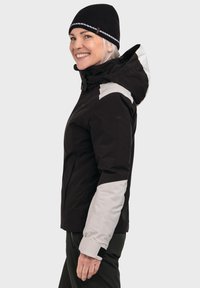 Schwarze Outdoor-Jacke mit grauen und weißen Akzenten, verstellbarer Kapuze und sicheren Bündchen. Das Material wirkt wasserdicht und langlebig, konzipiert für kaltes Wetter.