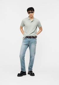 Άνδρας που φοράει ανοιχτό πράσινο πόλο πουκάμισο, ανοιχτό μπλε τζιν με κεντημένους αριθμούς, μαύρα χοντρά παπούτσια, σκούρα γυαλιά ηλίου, στέκεται με τα χέρια πίσω από την πλάτη.