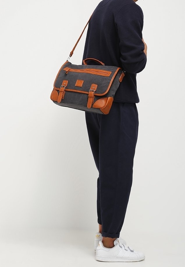 Personne portant une tenue bleu marine avec des baskets blanches, portant un sac messager en toile grise avec des sangles et des accents en cuir marron.
