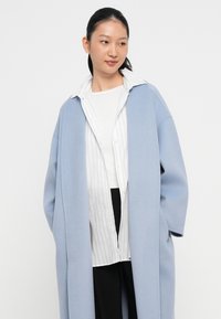 Abrigo de gran tamaño en un suave tono azul claro, confeccionado con una tela blanda, que presenta un cuello de chal y hombros caídos, usado sobre una camisa blanca a rayas y pantalones negros.