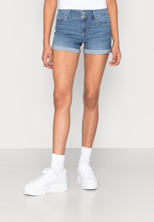 Femme portant un short en jean bleu retroussé, des chaussettes blanches et des baskets blanches à plateforme, debout devant un fond clair uni.