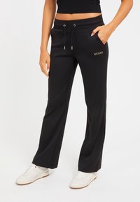 Kvinde iført sorte Guess sweatpants med snoretræk i taljen og hvide sneakers, stående foran en ensfarvet hvid baggrund.