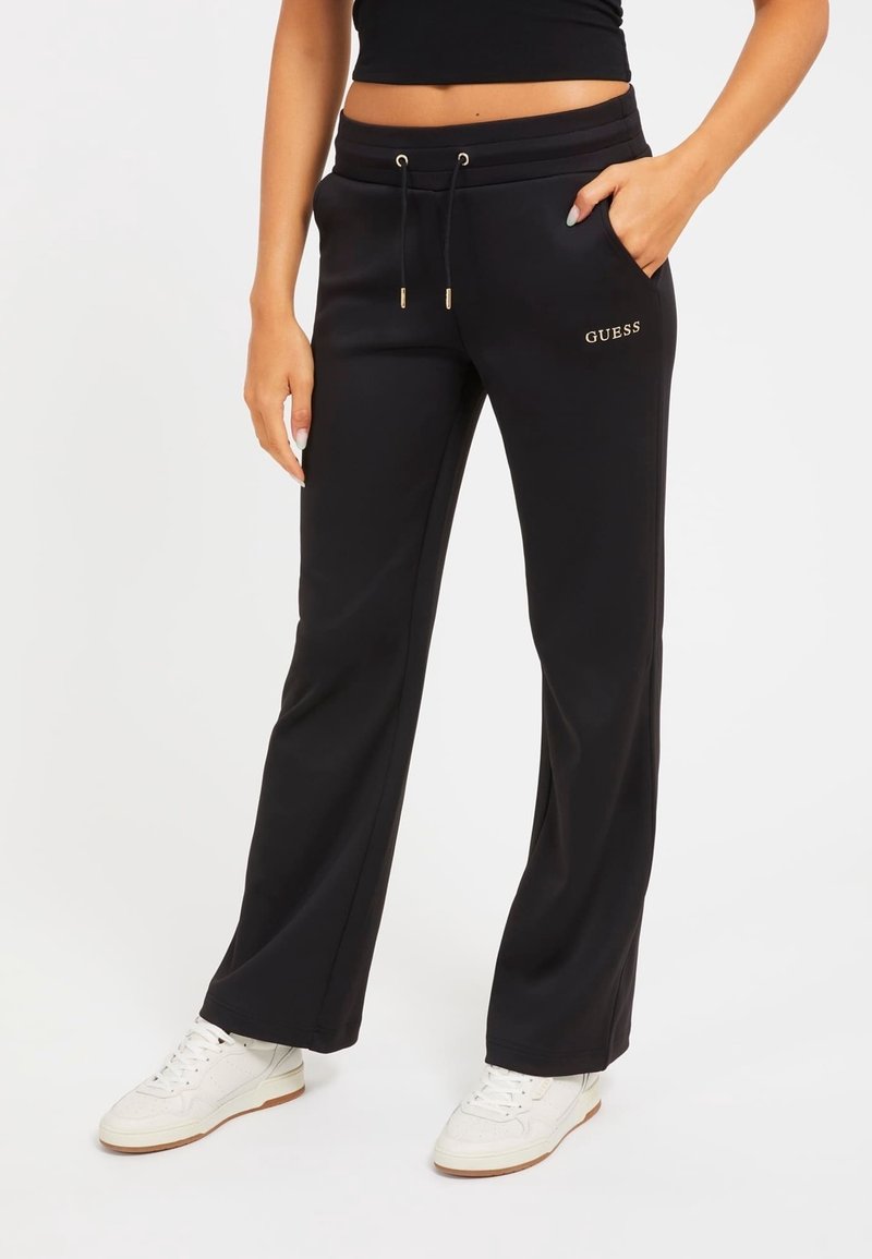 Kvinde iført sorte Guess sweatpants med snoretræk i taljen og hvide sneakers, stående foran en ensfarvet hvid baggrund.
