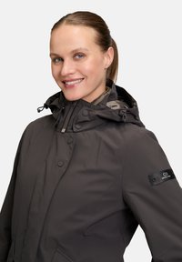 Dark gray waterproof jacket with a hood, snap button closure, and elastic drawstring. Features a logo patch on the left sleeve. Smooth texture.