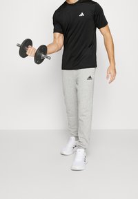 Camiseta deportiva negra con un logo blanco, pantalones de chándal grises, sosteniendo un dumbbell negro. Zapatillas deportivas blancas con detalles. Diseño simple y funcional.