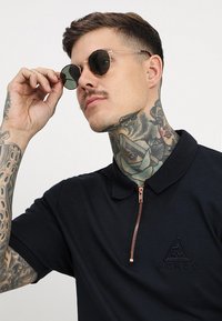 Cămașă polo neagră cu mânecă scurtă, guler cu fermoar, logo brodat și detalii cu fermoar cupru. Ochelari de soare cu lentile rotunde și tonificate.