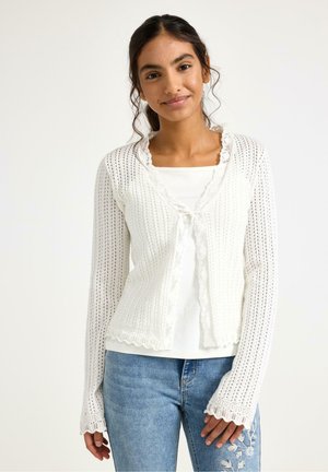 Ung kvinde iført en hvid, blondertrimmet strikket cardigan over en hvid top og broderede blå jeans, stående foran en ensfarvet baggrund.