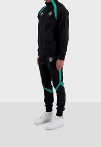 Černá tepláková souprava s tyrkysovými doplňky, zahrnující zipovou bundu a odpovídající kalhoty, obojí s logem; nošeno s bílými teniskami.