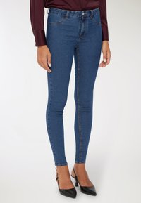 Kobieta w niebieskich wąskich dżinsach, burgundowej bluzce z długim rękawem i czarnych butach na obcasie z odkrytymi piętami, stojąca na tle białym.