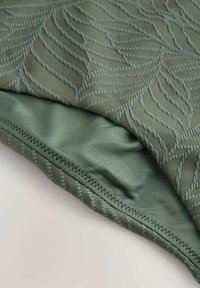 Material verde măsliniu cu un model texturat de frunze, cu broderie contrastantă în albastru deschis. Partea inferioară netedă, cu detalii vizibile de cusături.