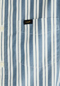 Chemise à rayures bleues et blanches avec une fermeture à boutons et une poche sur la poitrine gauche, comportant une étiquette noire avec "Lee®" en lettres dorées.