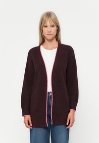 Cardigan bordeaux avec un tricot texturé, présentant une bordure rose le long de l'ouverture et des poignets des manches. Porté sur une chemise blanche et un jean.