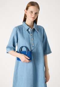 Denim-mekko lyhyillä hihoilla, suurella kauluksella ja napituslistalla; pitää käsissään pientä sinistä nahkalaukkua, jossa on pyöreät kahvat.