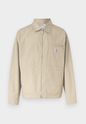 Chaqueta beige con cremallera y cuello, dos bolsillos delanteros y un pequeño parche con el logo de Carhartt en el bolsillo del pecho izquierdo.