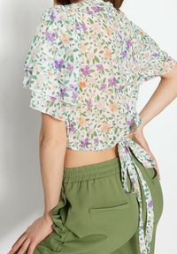 Top corto floreale in tessuto trasparente con maniche corte a volant, dotato di un fiocco sul retro. I colori includono rosa, viola, verde e arancione su uno sfondo chiaro.