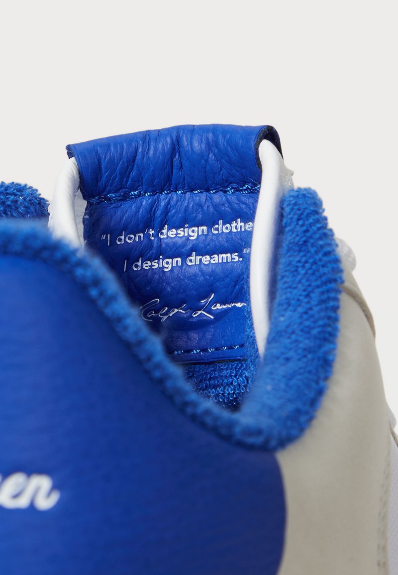 Weißer Sneaker mit blauen Textil- und Lederelementen. Das Innenfutter verfügt über eine gepolsterte blaue Zunge mit einem Zitat und einer Unterschrift in weißem Text.