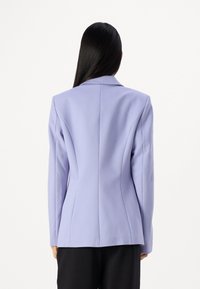 Persona con lunghi capelli neri indossa un blazer lavanda aderente e pantaloni neri, in piedi di spalle contro uno sfondo bianco semplice.