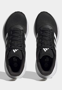 Zapatillas deportivas negras con parte superior texturizada, cordones negros, rayas blancas y suela blanca. Presentan el texto "FIN AL DESPERDICIO DE PLÁSTICO" en su interior.