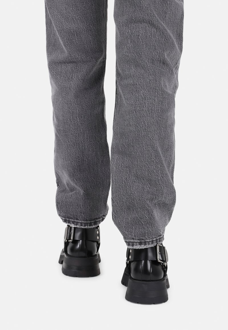 Graue Denim-Hosen von hinten betrachtet, mit einem lockeren Schnitt. Schwarze klobige Stiefel mit Schnallen-Detail und strukturierten Riemen sind sichtbar.