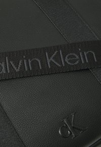 Schwarze Ledertasche mit strukturierten Oberflächen, einem auffälligen schwarzen Stoffgurt mit eingeprägtem "Calvin Klein" und einem dezenten eingeprägten Logo.