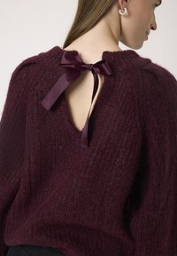Maglione lavorato a maglia color bordeaux con una texture a coste, caratterizzato da una schiena aperta fissata da un fiocco in raso scuro. Collo rotondo e maniche voluminose.