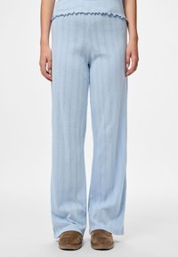 Ženska oblečena v svetlo modre pletene široke hlače in ujemajočo se bluzo ter rjave natikače, stoji ob ozadju iz čiste bele barve.