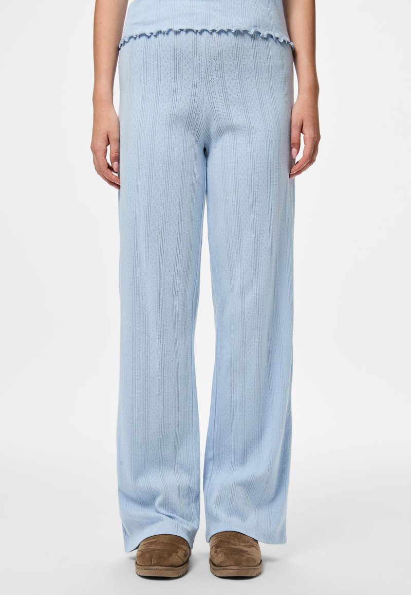 Ženska oblečena v svetlo modre pletene široke hlače in ujemajočo se bluzo ter rjave natikače, stoji ob ozadju iz čiste bele barve.