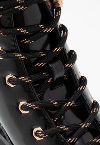 Botte en cuir verni noir avec des lacets noirs et dorés à motifs, dotée d'œillets en métal doré et d'une texture lisse et brillante.