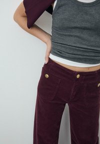 Szary prążkowany top na ramiączkach nałożony na biały podkoszulek, zestawiony z bordowymi sztruksowymi spodniami z dużymi złotymi guzikami i tylnymi kieszeniami.