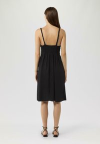 Schwarzes ärmelloses Kleid mit gerafftem Oberteil, fließendem Rock und dünnen Trägern. Begleitet von schwarzen Riemchensandalen. Glatter Stofftextur.