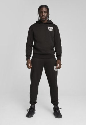 Hanorac negru cu glugă, dotat cu buzunar frontal și logo alb, asociat cu pantalonii de trening ajustați, care au logo-ul pe coapsa stângă.