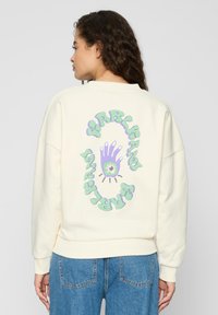 Krémová bavlnená mikina s voľným strihom. Na zadnej strane je farebná grafika ruky a štýlový text. Manžety a spodný lem sú ribované.