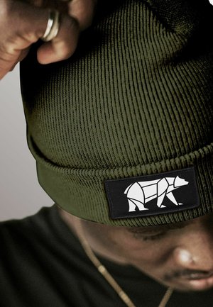 Person adjusting a green knit beanie with a black patch featuring a white geometric bear design.