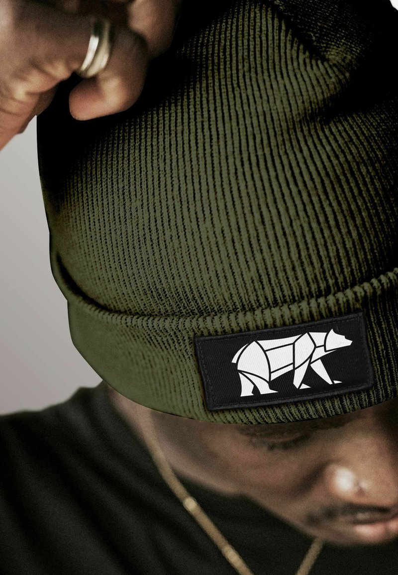 Person adjusting a green knit beanie with a black patch featuring a white geometric bear design.