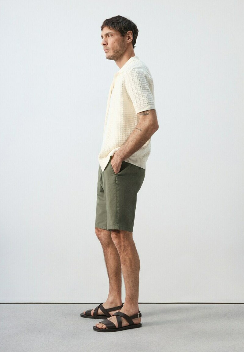 Homme debout de profil portant une chemise en tricot crème à manches courtes, un short vert olive et des sandales noires à brides croisées devant un fond uni.