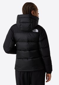 Persona con trenzas que lleva una chaqueta acolchada con capucha negra y el logo de The North Face, de espaldas contra un fondo sencillo.