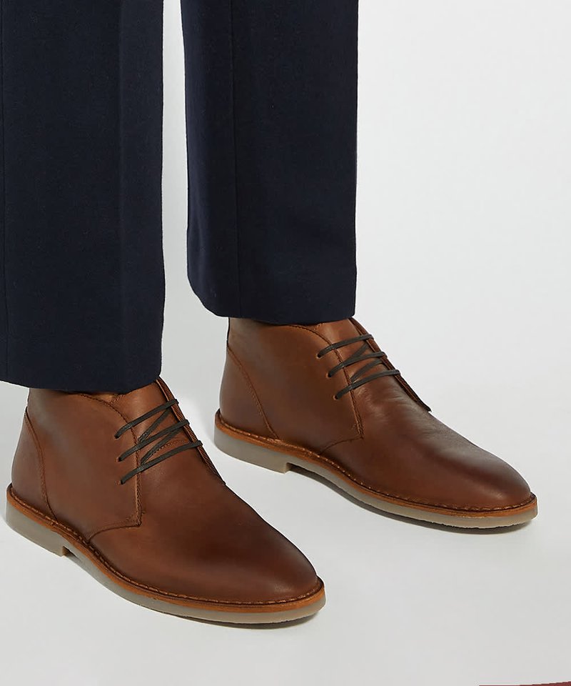 Bottines en cuir marron avec lacets foncés portées avec un pantalon bleu marine sur un fond clair uni.